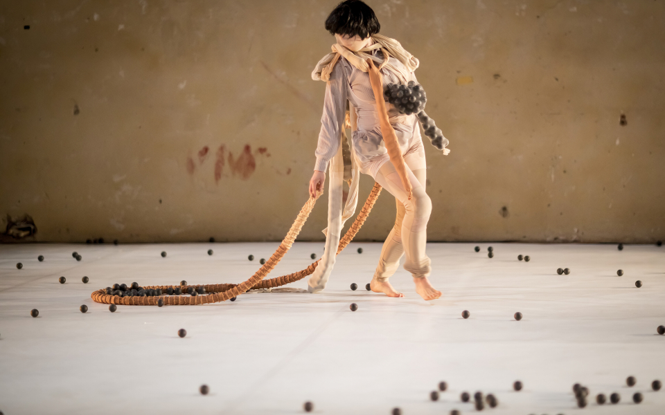 © Vojtech Brtnicky Eine Figur mit schwarzer Perücke, gekleidet in ein helles, dehnbares Tuch, läuft über eine Bühne, die mit kleinen schwarzen Bällen übersät ist. Launig zieht sie einen kleinen Haufen der schwarzen Bälle hinter sich her.