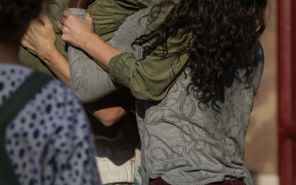 Katy is a white woman with long brown hair that falls down her back. She is wearing an olive green long-sleeved jumpsuit. Her head is tilted back and she sings energetically into a microphone she holds in her right hand while being carried on her shoulders by three other dancers. The wall of a sandstone-coloured house can be seen in the background.