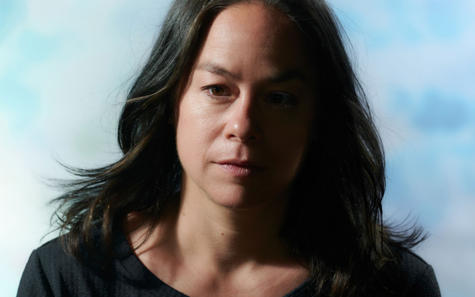 Portrait photo of Mirjam Gurtner against a light blue cloudy background. A white woman, with brown eyes and brown long hair worn loose, and a dark blue T-shirt.