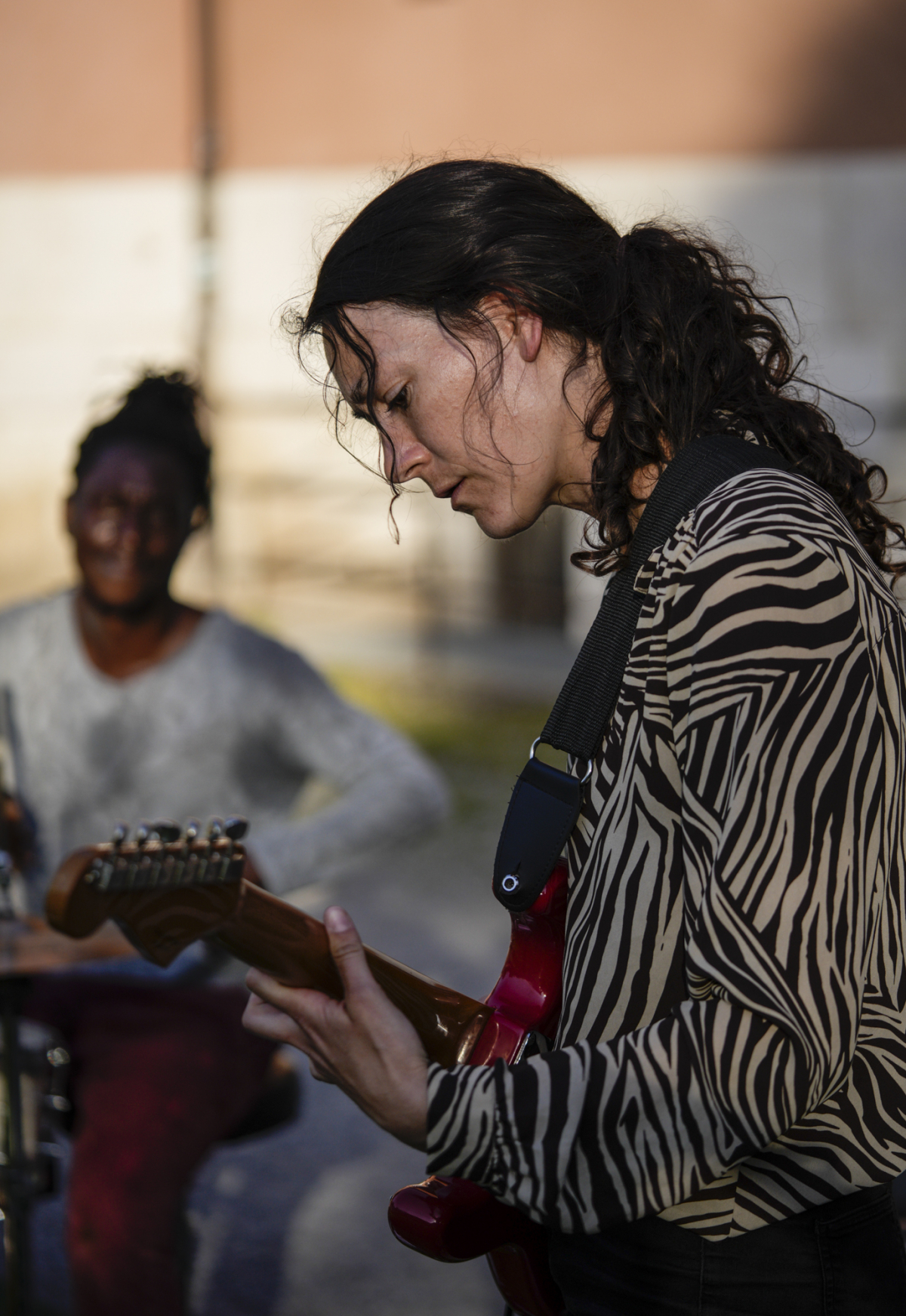 Teresa is a white woman with long brown hair tied back in a ponytail. She wears a black and white striped long-sleeved blouse. She plays a red guitar with a black neck strap. A dark-skinned second musician sits in the background and looks over at her.
